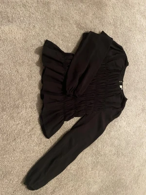 Svart långärmad blus från Nelly - Snygg svart blus med smockdetaljer över hela bröstet och midjan med volang nedtill. Blusen har rund halsringning och långa ärmar med lätt puff. Materialet är ett tunt, mjukt tyg som ger ett flowy intryck. Perfekt för dig som gillar stilrena plagg med lite extra detaljer. Är bara provad. Köpt för 299.