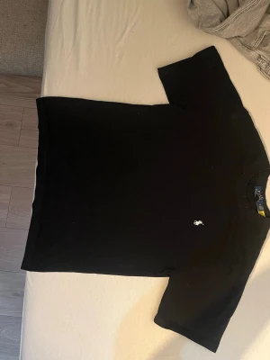 Svart t-shirt från Polo Ralph Lauren - Klassisk svart t-shirt från Polo Ralph Lauren med rund hals och den ikoniska vita broderade loggan på bröstet. Tillverkad i mjuk bomull och har en normal passform. Perfekt basplagg för en clean och stilren look.