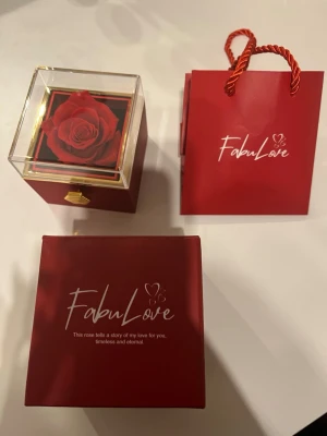 FabuLove smyckesbox med röd ros - Snygg smyckesbox från FabuLove i röd och guld med en konstgjord röd ros under ett genomskinligt lock. Perfekt för att förvara smycken eller som presentförpackning. Kommer med matchande röd presentpåse och elegant design.