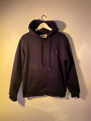 Svart hoodie Comme des Garçons Play - Svart hoodie från Comme des Garçons Play med dragkedja och snörning i huvan. Ikoniskt hjärta med ögon på bröstet och stort tryck på ryggen. Mjuk bomullskvalitet och praktisk känguruficka framtill. Perfekt för dig som gillar streetwear och snygga detaljer. Storlek S som även passar M