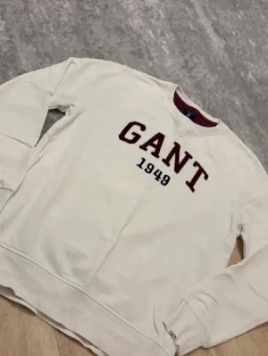Vit sweatshirt från GANT - Vit sweatshirt från GANT med vinröda bokstäver och siffran 1949 broderat på bröstet. Tröjan har rund halsringning med vinröd detalj på insidan, långa ärmar och ribbade muddar vid ärmslut och nederkant. Perfekt för en avslappnad och stilren look.