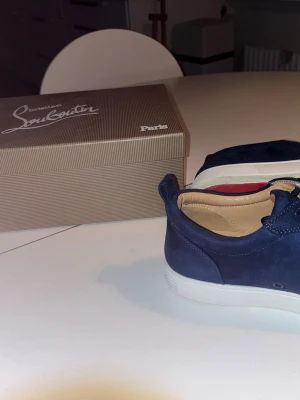 Blå sneakers från Christian Louboutin - Snygga mörkblå sneakers från Christian Louboutin i mocka med klassiska röda sulor och coola nitar framtill. Skorna har snörning och en låg profil som ger en modern look. Perfekta för dig som vill sticka ut med exklusiva detaljer. Bytt någon nit men annars perfekt skick. Allt og förutom en spik 