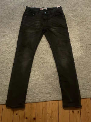 Svarta/gråa Levi's 512 - Säljer ett par svarta Levi's 512 slim jeans med klassisk femficksdesign och knappgylf. Jeansen har en fet passform och är tillverkade i mjukt bomullsmaterial med lite stretch för extra komfort. Storleken är 14a barn men dom sitter som 28/30 i herrstorlek ända defekten är lappen bak som har slitits 