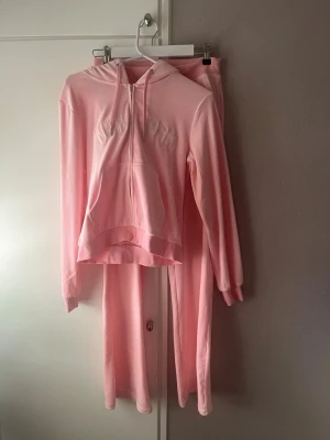 Mjukisset  - Supermysigt ljusrosa loungewear-set med hoodie och matchande byxor. Hoodien har dragkedja, ficka framtill och broderad text. Byxorna har raka ben, dragsko i midjan och fickor. Perfekt för chill dagar hemma.