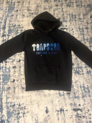 Svart hoodie från Trapstar med tryck - Säljer en svart hoodie från Trapstar med blått och vitt tryck på bröstet. Hoodien har en klassisk huva med dragsko och en stor magficka framtill. Perfekt för dig som gillar streetwear och vill ha en snygg och bekväm stil.
