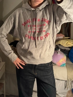 Grå Zadig & Voltaire hoodie med tryck - Snygg grå hoodie från Zadig & Voltaire med rött tryck framtill och glittriga detaljer. Klassisk känguruficka och justerbar huva med snören. Perfekt för en avslappnad och trendig look.