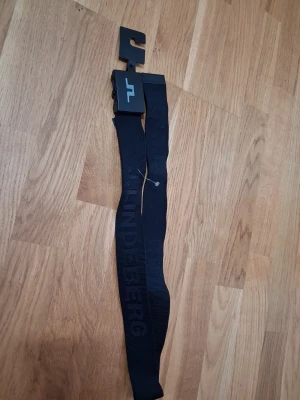 Svart tygbälte från J.Lindeberg - Snyggt svart tygbälte från J.Lindeberg med logga på metallspännet och diskret text längs bandet. Bältet är 85 cm långt och har en modern, sportig look som passar till jeans eller chinos.