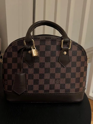  Alma handväska -  Väskan har guldfärgade metalldetaljer, dubbla handtag och en hängande tag. Tillverkad i canvas med detaljer i mörkbrunt skinn. Perfekt för dig som vill ha en ikonisk och stilren accessoar.