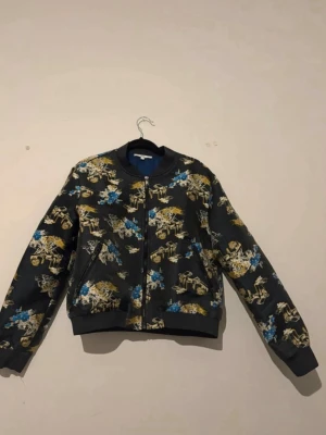Mönstrad jacka - Köpt i Frankrike för 1200kr, passar både S och XS Unik bomberjacka med svart bas och blommigt mönster i guld, blått och vitt. Jackan har klassisk bomberkrage, ribbade muddar och dragkedja framtill. Perfekt för dig som vill sticka ut med en cool och färgstark look. 