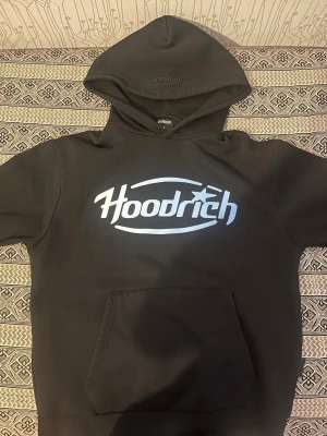 Svart hoodie från Hoodrich - [Byxorna finns på min profil.] Svart hoodie från Hoodrich med stor vit logga på bröstet och broderad logga på huvan. Klassisk känguruficka framtill och mjukt material som känns skönt mot huden. Perfekt för en avslappnad streetwear-look.