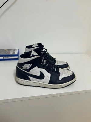 Nike Air Jordan 1 svartvita sneakers - Säljer ett par klassiska Nike Air Jordan 1 sneakers i svart och vitt. Skorna har hög siluett, snörning och den ikoniska Air Jordan-loggan på sidan. Tillverkade i skinn med svart swoosh och vit bas. Perfekta för dig som gillar streetwear och tidlös stil.