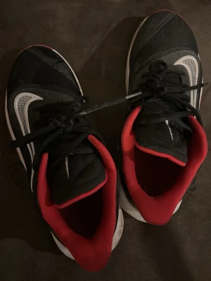 Svarta Nike sneakers med röd detalj - Säljer ett par svarta Nike sneakers med vit sula och vit swoosh på sidan. Skorna har röda detaljer på insidan och svarta skosnören. Ovandelen är i textil och syntetmaterial, designade för en sportig och modern look.