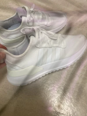 Vita adidas sneakers med snörning - Snygga helvita adidas sneakers med klassiska tre ränder på sidan. Skorna har snörning och en låg profil med rund tå. Ovandelen är i mesh och syntetmaterial för en sportig look. Perfekta för dig som gillar stilrena och bekväma sneakers.