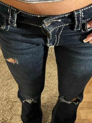Mörkblå lågmidjade jeans med slitningar - Mörkblå jeans med vita kontrastsömmar och slitningar vid knä och benslut. Lågmidjad modell och klassiska fickor bak med lock och knapp. Perfekta för en trendig och avslappnad look.