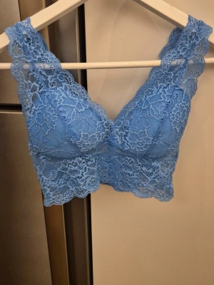 Blå spetsbralette med breda axelband - Snygg blå bralette i spets med breda axelband och vacker blommig spetsdetalj. Bh:n har vadderade kupor och längre spetskant nedtill för extra stöd och stil. Perfekt för dig som gillar en feminin och bekväm look.
