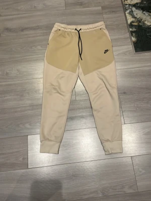 Beige sweatpants från Nike - Snygga beige sweatpants från Nike med tvåfärgad design och svart logga på vänster sida. Byxorna har resår i midjan med svart dragsko och muddar vid bensluten. Perfekta för träning eller chill. Tillverkade i mjukt bomullsmaterial.