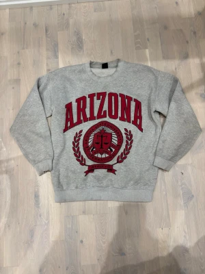 Grå sweatshirt med Arizona-tryck - Snygg grå sweatshirt med stort rött Arizona-tryck och emblem framtill. Tröjan har rund hals, långa ärmar och ribbade muddar. Perfekt för en avslappnad och sportig stil. Mjuk bomullskänsla och klassisk passform.
