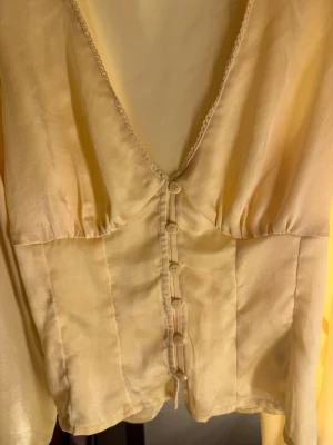 Ljusgul satinblus med knappar - Supersöt ljusgul blus i satinliknande material med djup v-ringning, dekorativa knappar framtill och knytband i ryggen. Blusen har långa ärmar och lätt puff vid axlarna 