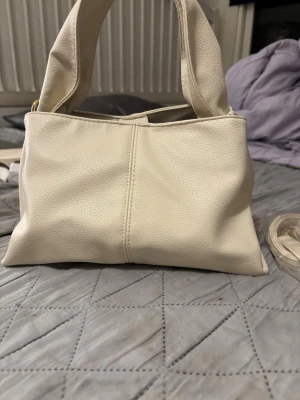 Krämvit handväska i skinnimitation - Stilren krämvit handväska med mjuka former och breda handtag. Väskan är tillverkad i skinnimitation och har ett magnetlås. Insidan är fodrad i vinrött tyg. Perfekt storlek för det viktigaste och enkel att matcha till olika outfits.