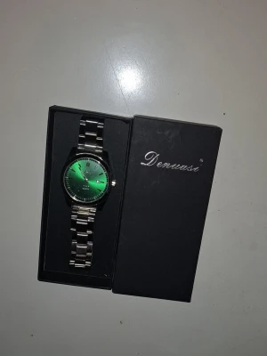 Stilren Denwase klocka i grön urtavla - Säljer nu denna helt nya Denwase klocka, med en stilren grön urtavla. Klockan passar till alla outfits och är perfekt för dig som vill sticka ut lite i vardagen med lyx samt en prisvärd klocka. Box medföljer, priset är självklart diskuterbart 🙂 länkar medföljer