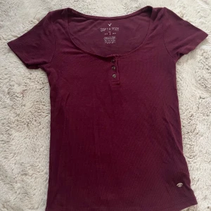 Vinröd ribbad topp från American Eagle - Superskön vinröd ribbad topp från American Eagle med rundad halsringning och tre knappar framtill. Kortärmad och figurnära passform, perfekt för dig som gillar enkel men snygg stil. Soft & Sexy-kollektionen, stretchigt bomullsmaterial.