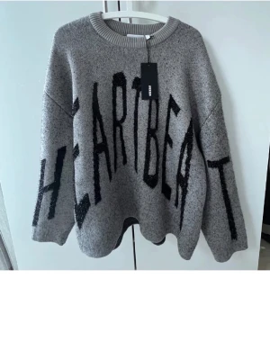 Grå stickad tröja med text - Oversized grå stickad tröja med svart texten 'HEARTBEAT' framtill. Tröjan har rund halsringning och långa ärmar. Perfekt för dig som gillar statement-plagg och en avslappnad stil. Säljer då den är för stor på mig 🥰