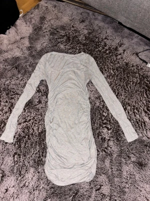 Ljusgrå långärmad klänning - En enkel och stilren ljusgrå klänning med långa ärmar och rund halsringning. Klänningen är figurnära och har rynkade detaljer längs sidorna för en snygg siluett. Perfekt att styla med sneakers eller boots.