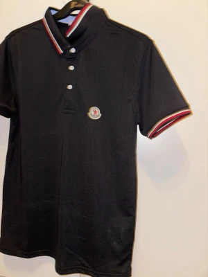 Svart Moncler pikétröja med röd/vita detaljer - Klassisk svart pikétröja från Moncler med korta ärmar, vit och röd rand på krage och ärmslut samt broderad Moncler-logga på bröstet. Tröjan har knappar framtill och är tillverkad i mjuk bomull.