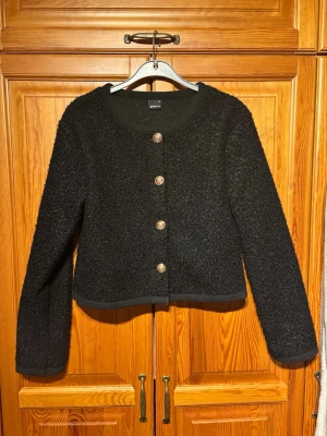 Svart kofta med guldknappar - Stilren svart kofta från Gina Tricot med rund halsringning och fyra dekorativa guldknappar framtill. Koftan har lång ärm och är tillverkad i ett mjukt, lite fluffigt material som ger en lyxig känsla. Perfekt att styla till både jeans och kjol. Använd 1 gång!