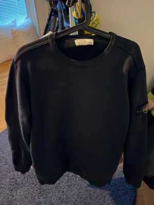 Svart sweatshirt från Stone Island - Klassisk svart sweatshirt från Stone Island med rund halsringning och ikonisk logotyp-patch på vänster ärm. Tröjan har en stilren design och är tillverkad i mjukt material som passar perfekt till streetwear-stilen.