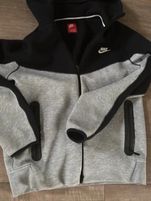 Nike hoodie i svart och grå, M - Nike hoodie i svart och ljusgrått med dragkedja framtill och två fickor med svarta detaljer. Klassisk Nike-logga på bröstet och huva. Perfekt för en sportig och avslappnad stil.