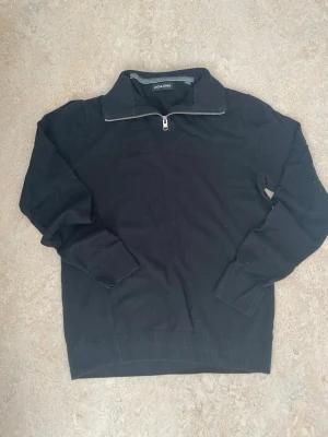 Svart stickad tröja med halv dragkedja - Svart stickad tröja från Jack & Jones med hög krage och halv dragkedja. Tröjan har ribbade muddar vid ärmslut och nederkant samt en stilren look som passar till det mesta. Perfekt för lager på lager och enkel att matcha.