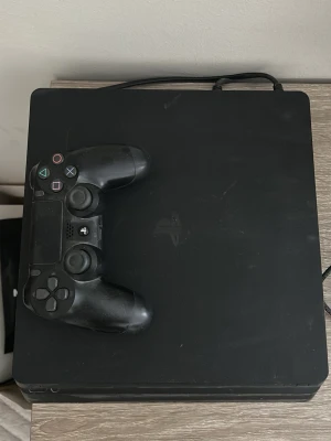 PlayStation 4 Slim med handkontroll - Säljer en svart PlayStation 4 Slim med tillhörande svart DualShock 4-handkontroll. Konsolen har en stilren, fyrkantig design och är tillverkad i plast. Perfekt för gaming och streaming av film och serier.