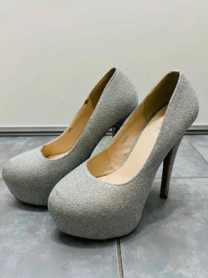 Glittriga silverfärgade pumps - Snygga pumps i glittrigt silver från Fashion Ye. Skorna har rund tå, hög klack och plattform för extra höjd. Perfekta för dig som vill sticka ut med ett par riktigt glamorösa klackar. Insidan är beige och materialet känns syntetiskt.