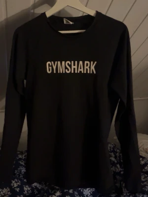 Svart långärmad tröja Gymshark - Svart långärmad tröja från Gymshark med vit logga tryckt på bröstet. Tröjan har rund halsringning och är tillverkad i ett mjukt och stretchigt material som passar perfekt till träning eller chill. Enkel och stilren design.