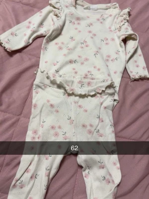 Blommigt babyset med volangdetaljer - Sött tvådelat set för baby i mjuk bomull, med långärmad body och matchande byxor. Setet är vitt med ljusrosa blommönster och har volangdetaljer vid axlar och midja. Perfekt för dig som gillar gulliga och bekväma plagg till de minsta.