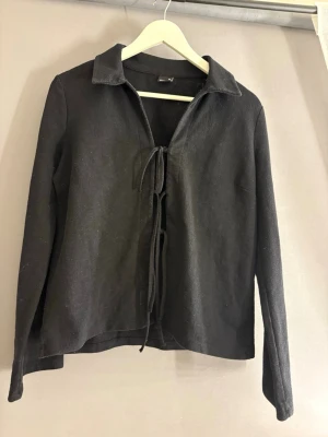 Svart långärmad tröja med knytning - Svart långärmad tröja med krage och snygg knytning framtill. Tröjan har en rak passform och är tillverkad i ett mjukt tyg som känns bekvämt mot huden. Perfekt för en stilren och trendig look.