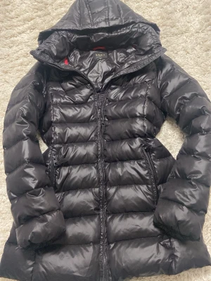 SAKI Jacka! - Snygg svart dunjacka från Saki med quiltad design och huva. Jackan har dragkedja framtill och två sidofickor. Perfekt för kalla dagar och ger en clean, modern look. Org pris mellan 2000-3000kr!!