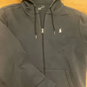 Svart hoodie från Polo Ralph Lauren - Snygg svart hoodie med dragkedja från Polo Ralph Lauren. Klassisk design med vit broderad logga på bröstet, justerbar huva med snören och fickor framtill. Perfekt för en avslappnad och stilren look. Den är solblekt
