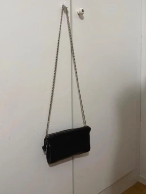 Svart axelväska med kedjerem - Snygg svart axelväska med stilren silverfärgad kedjerem och dragkedja upptill. Väskan har en avlång, minimalistisk form och är tillverkad i slätt skinnliknande material. Perfekt för dig som gillar enkel men trendig design.