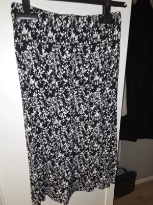 Svartvit blommig långkjol - Snygg svart långkjol med vitt blommönster. Kjolen har en rak och flowy passform som ger en avslappnad men ändå stilren look. Perfekt att matcha med en enkel topp eller stickad tröja. Materialet känns mjukt och följsamt.