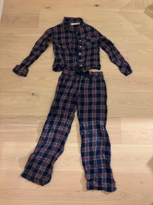 Rutig pyjamas från Gina Tricot - Mysig tvådelad pyjamas från Gina Tricot i klassiskt rutigt mönster med mörkblå, röda och gröna toner. Skjortan har knappar framtill och två bröstfickor, byxorna har resår i midjan. Perfekt för chillkvällar hemma.