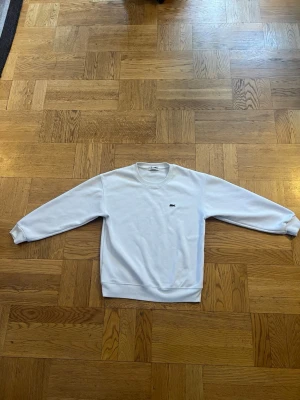 Vit sweatshirt från Lacoste - Klassisk vit sweatshirt från Lacoste med rund halsringning och den ikoniska gröna krokodilloggan broderad på bröstet. Tröjan har ribbade muddar vid ärmslut och nederkant. Perfekt för en clean och stilren look. 