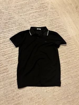 Svart pikétröja från Lacoste - Klassisk svart pikétröja från Lacoste med vit kant på kragen och ärmsluten. Tröjan har korta ärmar, knappslå framtill och den ikoniska krokodilloggan på bröstet. Perfekt till jeans eller shorts för en clean look.