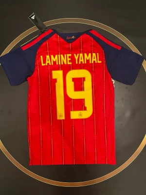 Spanien Lamine Yamal matchtröja - Röd och marinblå Spanien fotbollströja från Adidas med Lamine Yamal och nummer 19 på ryggen. Tröjan har gula detaljer, korta ärmar och klubbmärke på bröstet. Perfekt för dig som vill stötta landslaget på matchdag!