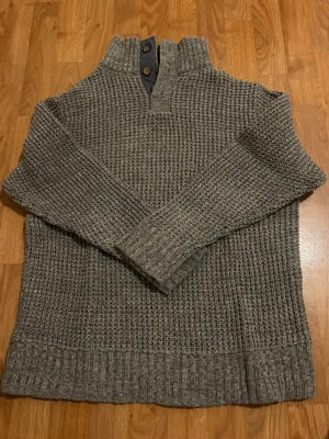 Grå stickad tröja med knappar - Stickad grå tröja med hög krage och två dekorativa knappar vid halsen. Grovstickat mönster och ribbade muddar vid ärmslut och nederkant. Perfekt för kyliga dagar och enkel att matcha med jeans.