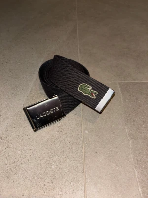 Svart tygbälte från Lacoste - Stilrent svart tygbälte från Lacoste med klassisk silverfärgad spänne och broderad grön krokodil-logga. Perfekt accessoar för att lyfta din outfit med en sportig touch. Justerbar längd och enkel att matcha med olika stilar.