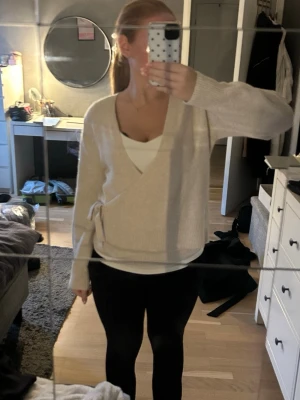 Beige omlottstickad tröja - Beige stickad omlott-tröja med knyt i sidan och v-ringad hals. Tröjan har långa ärmar och en mjuk, avslappnad passform. Perfekt att styla med ett linne under för en chill look.