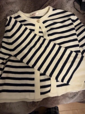Randig stickad cardigan i vitt och marinblått - Säljer en snygg stickad cardigan med vita och marinblå ränder. Tröjan har rund halsringning, långa ärmar och knäppning med två knappar framtill. Perfekt att slänga över ett linne för en chill och trendig look.