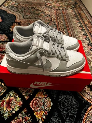 Nike Dunk Low vit/grå sneakers stolek 43 - Nike Dunk Low sneakers i vitt och ljusgrått med klassisk siluett. Skorna har snörning, rund tå och platt sula. Tillverkade i syntet och skinn med Nike-logga på sidan och hälen. Perfekta för dig som gillar stilrena och fräscha sneakers.